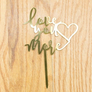 May include: A gold-colored cake topper with the words "love you more" in a cursive font, accompanied by a heart shape. The topper is made of a shiny material and is set against a wooden background.
