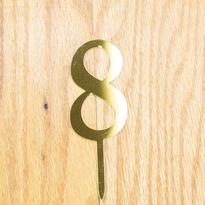 Op de afbeelding: Gouden taarttopper met het nummer acht en een dunne, gouden steel. De acht is een eenvoudig, modern ontwerp.