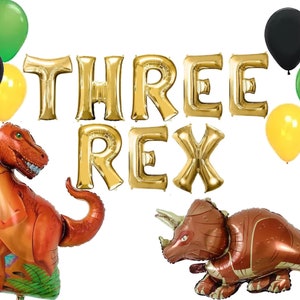 May include: Gold foil balloons spelling "THREE REX" with green, yellow, and black balloons.  There are also two dinosaur shaped balloons, one is a T-Rex and the other is a Triceratops.