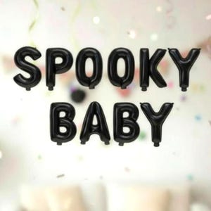 May include: Black foil balloons spell out "SPOOKY BABY" against a blurred background with confetti. The balloons are inflated and arranged in two rows. The theme is Halloween or a spooky baby shower.