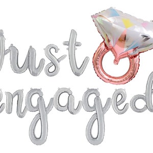 以下が含まれることがあります： 「just engaged」と綴られた銀色のフォイルバルーンレターと、ダイヤモンド型のトップが付いたローズゴールドのリングバルーン。