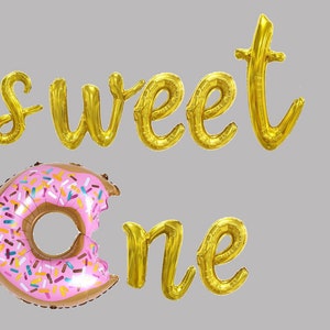 May include: Gold foil balloons spelling "sweet one" with a pink and brown donut balloon with colorful sprinkles.