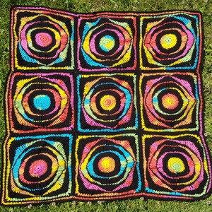 May include: A handmade, square crochet blanket with a vibrant, colorful design. The blanket features nine black squares, each with a central circle of rainbow-colored concentric rings. The blanket is made of soft yarn and is perfect for adding a pop of color to any room.