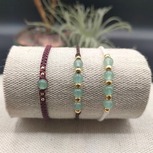 Peut inclure: Trois bracelets avec des perles de pierres précieuses vertes et des accents dorés. Les bracelets sont sur une surface en tissu beige. Les bracelets sont faits de ficelle rouge, blanche et marron.