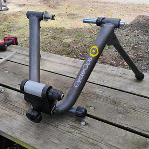 May include: A gray CycleOps bicycle trainer stand featuring a metal frame, a rear wheel mounting mechanism with a light gray roller, and a black adjustment knob. The brand name CycleOps is printed in white text with a yellow circular logo on the frame.