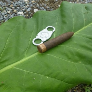 Pode incluir: Um charuto castanho repousando sobre uma grande folha verde com um cortador de charutos prateado ao lado.