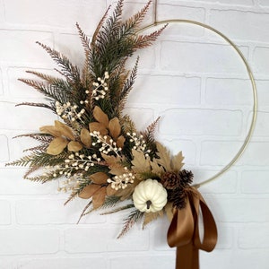 May include: A fall-themed wreath with a gold metal ring base. The wreath features faux greenery, beige leaves, white berries, a small white pumpkin, and pinecones. A brown ribbon is tied at the bottom. The wreath is hanging against a white brick wall.