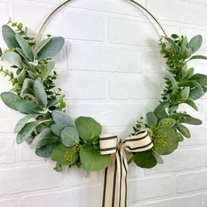 May include: A gold metal wreath with green eucalyptus leaves and a black and white striped ribbon.