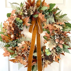 May include: A circular wreath featuring a dense arrangement of brown and orange autumn leaves, green fern fronds, pinecones, and small round seed pods. A long, smooth, dark mustard-colored velvet ribbon hangs vertically from the top center.