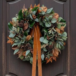 May include: A circular wreath featuring green eucalyptus leaves, brown dried foliage, and pinecones, accented with a long, hanging velvet ribbon in a warm amber tone. Below, a terracotta pot holds dried leaves and small pumpkins beside brown boots.