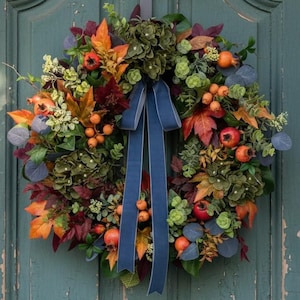 Puede incluir: Una corona de otoño circular que presenta una mezcla de follaje verde, naranja y rojo oscuro, pequeñas bayas naranjas y flores de hortensia verde oscuro. Una cinta larga de color azul marino cuelga del centro sobre una puerta verde.