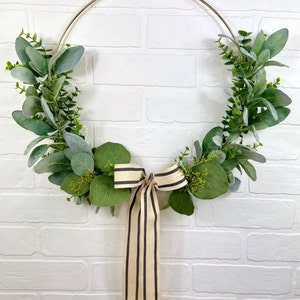 May include: A gold metal hoop wreath decorated with green eucalyptus leaves and a black and white striped ribbon bow.
