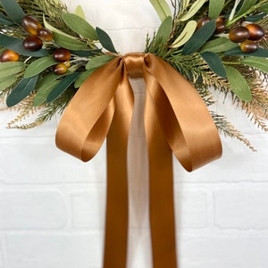 May include: A brown satin ribbon tied into a bow, hanging from a green and brown wreath. The wreath is made of artificial greenery and berries.