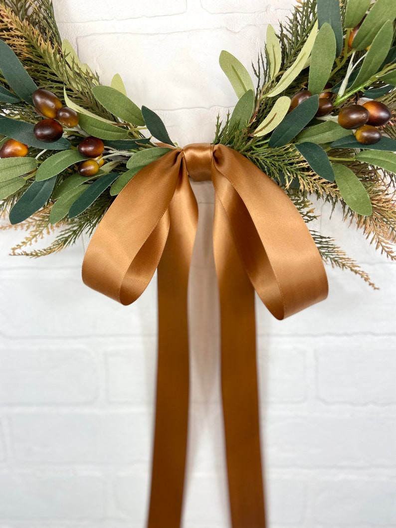 May include: A brown satin ribbon tied into a bow, hanging from a green and brown wreath. The wreath is made of artificial greenery and berries.