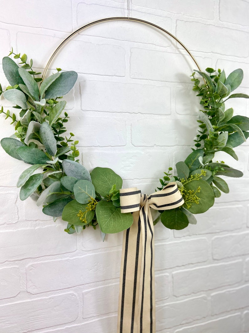 May include: A gold metal wreath with green eucalyptus leaves and a black and white striped ribbon.