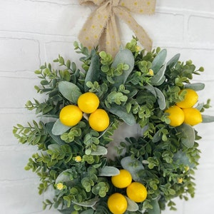 May include: A green and gray wreath with yellow lemons and a yellow polka dot bow. The wreath is hanging on a white brick wall.