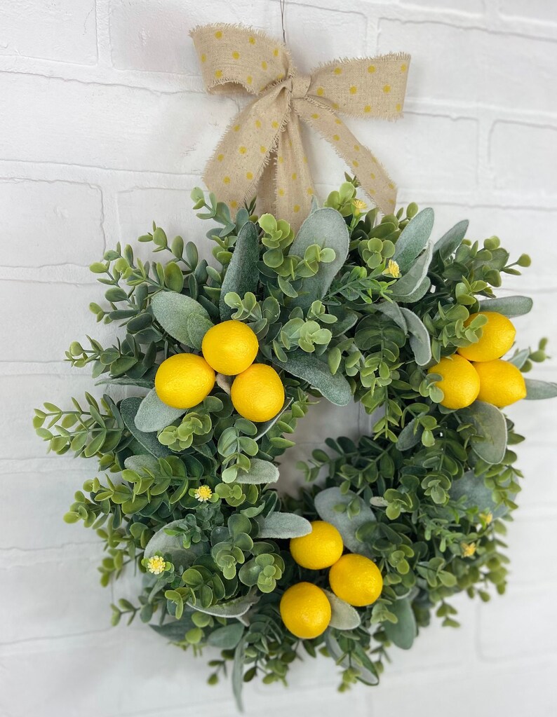 May include: A green and gray wreath with yellow lemons and a yellow polka dot bow. The wreath is hanging on a white brick wall.