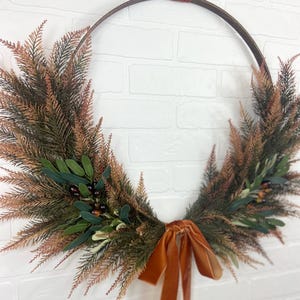 May include: A brown metal wreath with a burnt orange velvet ribbon. The wreath is decorated with faux greenery and berries.
