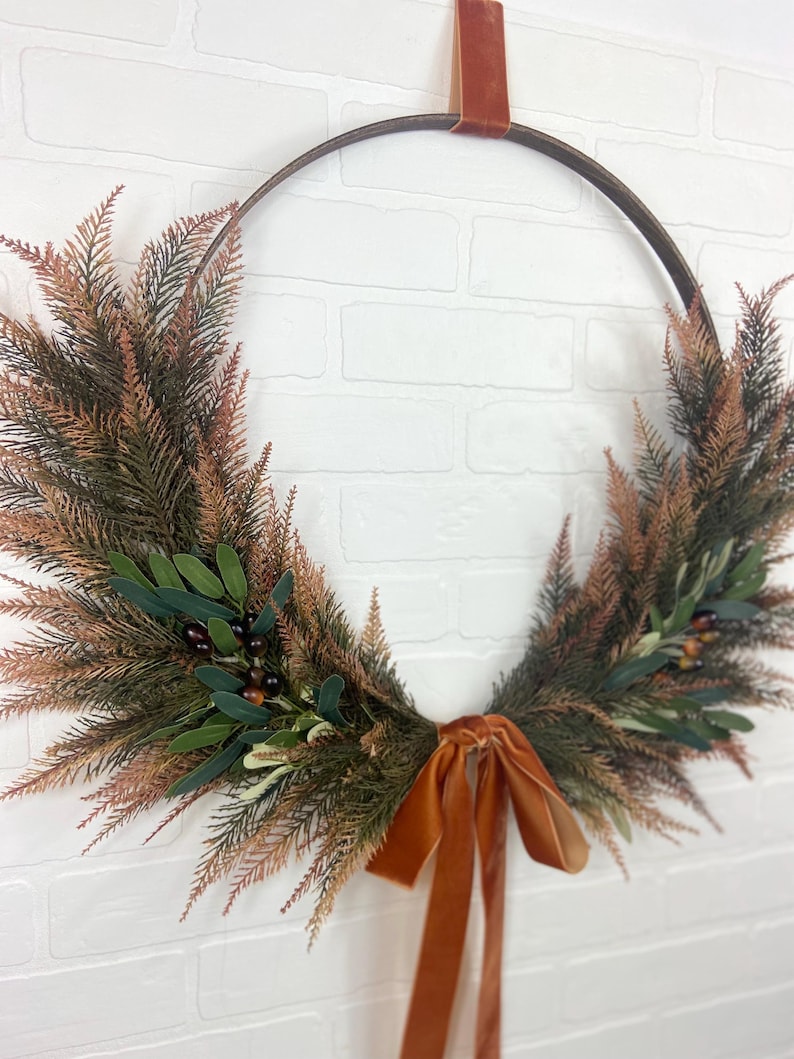 May include: A brown metal wreath with a burnt orange velvet ribbon. The wreath is decorated with faux greenery and berries.