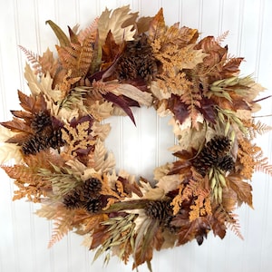 May include: A circular autumn wreath featuring a dense arrangement of faux maple leaves in shades of cream, tan, and deep brown. The design incorporates dark brown pinecones, textured orange-brown fern fronds, and slender stalks of dried wheat.