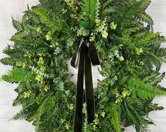 May include: A green fern wreath with a dark green velvet ribbon. The wreath is made of artificial greenery and is perfect for a spring or summer decor.