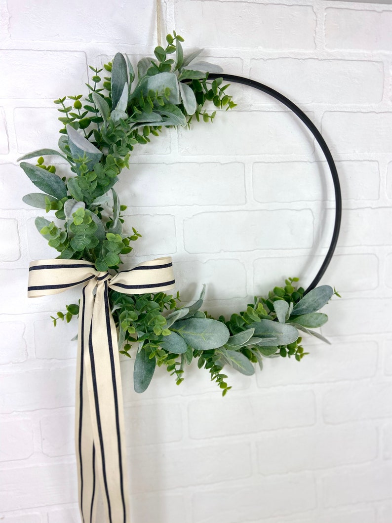 May include: A black metal wreath with a green and gray eucalyptus garland and a black and white striped ribbon.