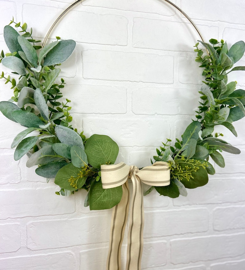 May include: A gold metal wreath with green eucalyptus leaves and a beige and brown striped ribbon bow.