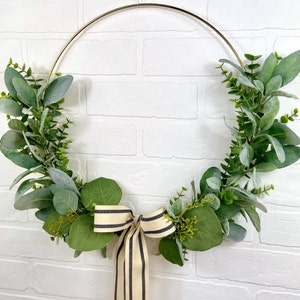 May include: A gold metal hoop wreath decorated with green eucalyptus leaves and a black and white striped ribbon bow.