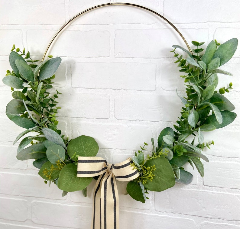 May include: A gold metal hoop wreath decorated with green eucalyptus leaves and a black and white striped ribbon bow.
