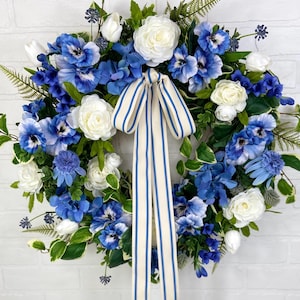 May include: A blue and white floral wreath with a white and blue striped ribbon. The wreath is made of artificial flowers and greenery.