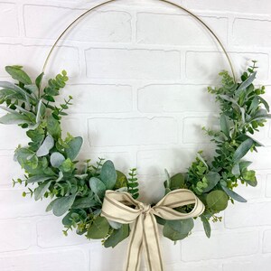 May include: A gold metal wreath with a cream and brown striped ribbon bow. The wreath is decorated with green and gray eucalyptus leaves.