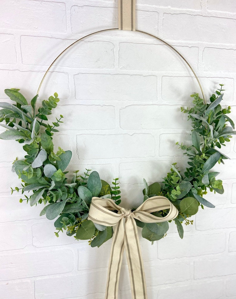 May include: A gold metal wreath with a cream and brown striped ribbon bow. The wreath is decorated with green and gray eucalyptus leaves.