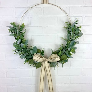 May include: A gold metal hoop wreath decorated with green eucalyptus leaves and a beige and brown striped ribbon bow.