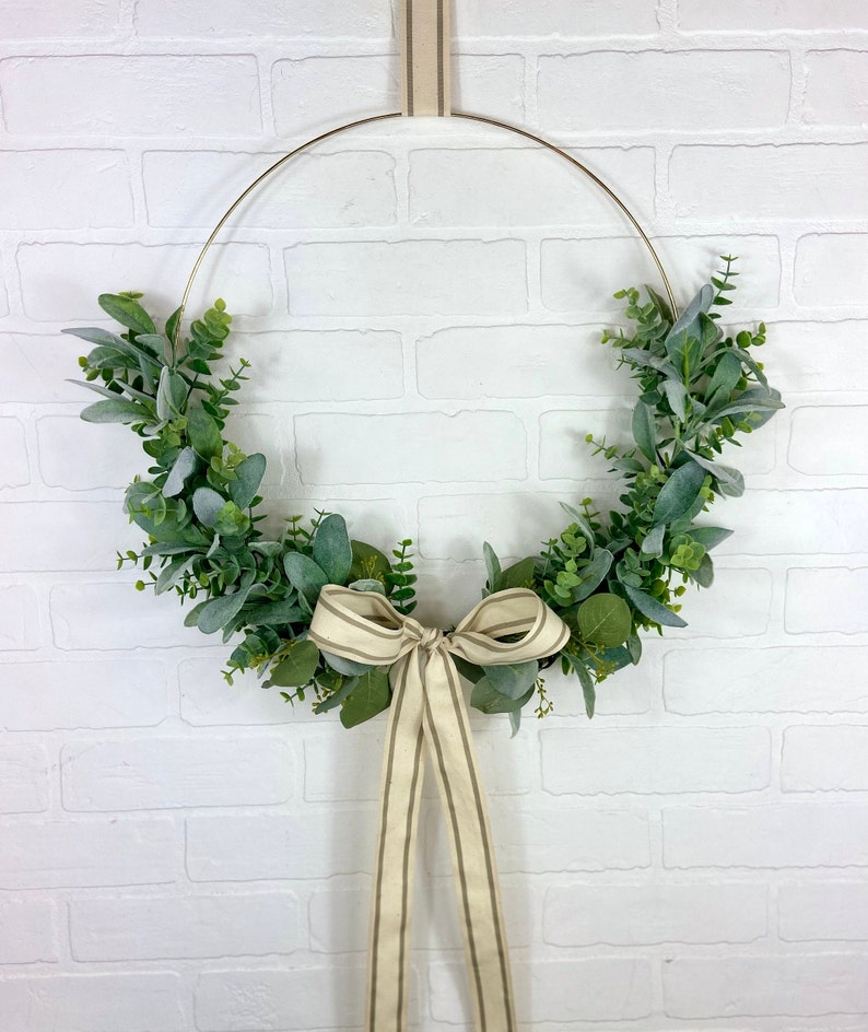 May include: A gold metal hoop wreath decorated with green eucalyptus leaves and a beige and brown striped ribbon bow.