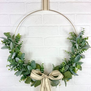 May include: A gold metal wreath with a green and white eucalyptus garland and a beige and brown striped ribbon bow.