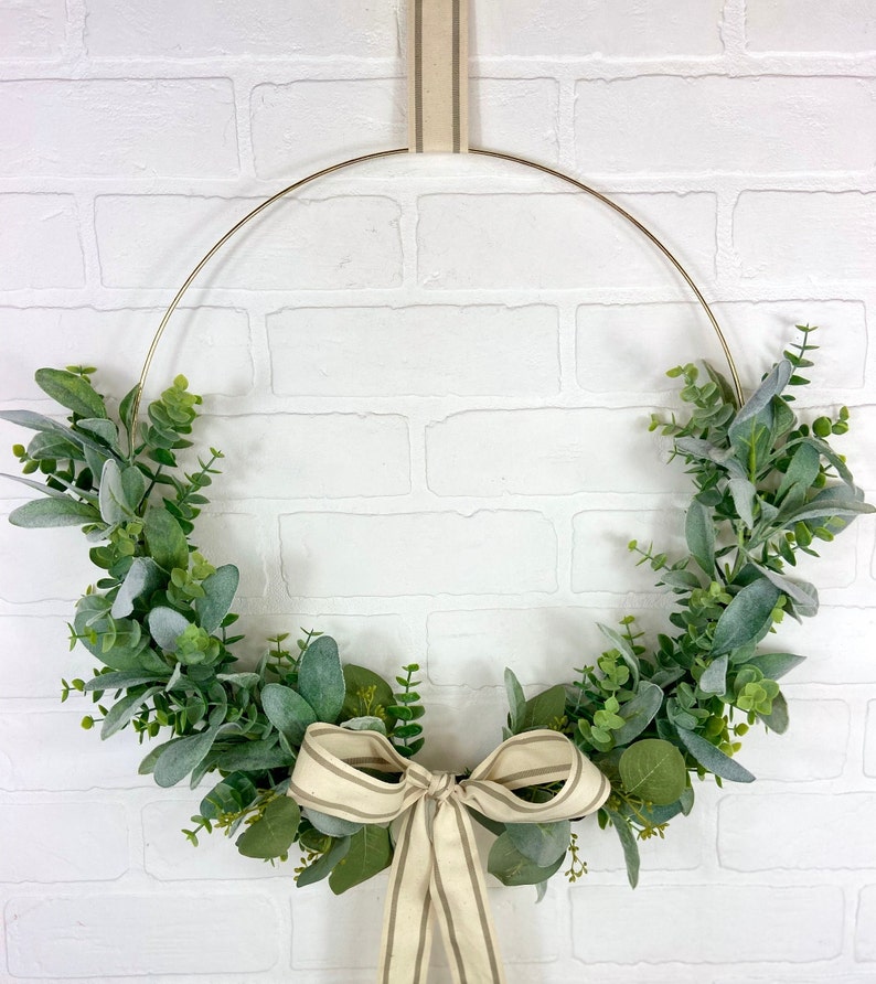 May include: A gold metal wreath with a green and white eucalyptus garland and a beige and brown striped ribbon bow.