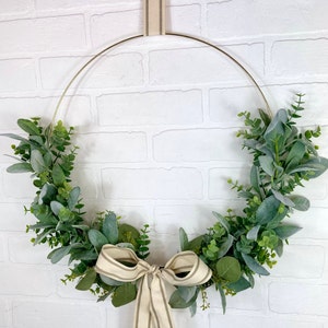 May include: A gold metal wreath with a cream and brown striped ribbon bow. The wreath is decorated with green eucalyptus leaves.