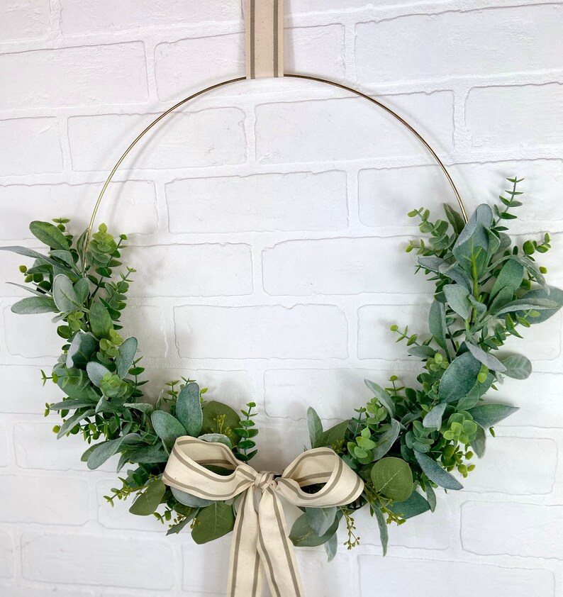 May include: A gold metal wreath with a cream and brown striped ribbon bow. The wreath is decorated with green eucalyptus leaves.