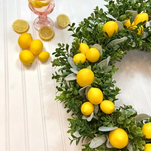 May include: A lemon wreath with faux lemons and greenery. The wreath is on a white background with a glass of lemonade and lemons.