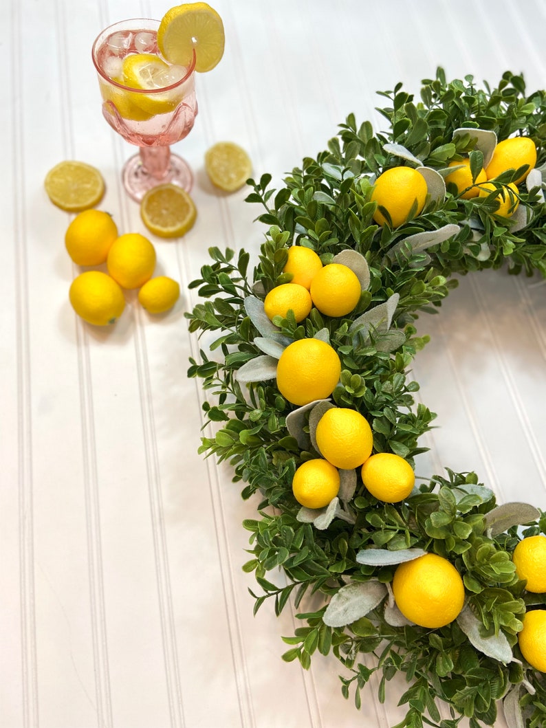 May include: A lemon wreath with faux lemons and greenery. The wreath is on a white background with a glass of lemonade and lemons.