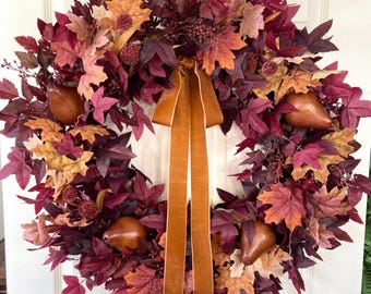 Könnte beinhalten: Ein herbstliches Kranz mit Blättern in Burgunder-, Orange- und Gelbtönen. Der Kranz hat künstliche Birnen und ein breites, goldbraunes Band. Dieser dekorative Kranz ist ideal für die Herbstsaison.