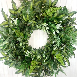 May include: A lush, green wreath composed of various faux foliage, including ferns, olive branches, and eucalyptus. Dark berries are interspersed throughout the wreath, adding depth and contrast. The wreath is circular and appears to be suitable for door or wall decor.