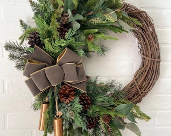 May include: A festive Christmas wreath featuring a woven grapevine base, adorned with faux pine and eucalyptus greenery, pinecones, and a brown bow with gold trim. Includes copper-colored bells for added charm.