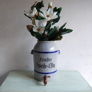 Könnte beinhalten: Eine weiße Keramik-Kanne mit blauem Besatz und einem Holzhahn. Die Kanne trägt den Schriftzug "Feinster Speise-Essig". Die Kanne ist mit weißen Blumen gefüllt.