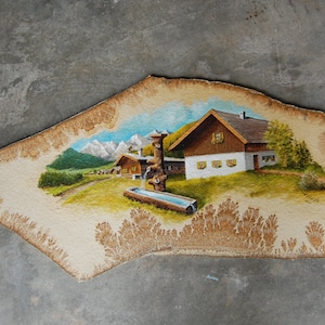 Puede incluir: Una escena pintada de un pequeño pueblo en las montañas. La pintura está sobre una pieza de madera con una superficie rugosa y texturizada. El pueblo está formado por unas pocas casas pequeñas con techos marrones y paredes blancas. Hay una pequeña fuente en primer plano con un abrevadero de madera. Las montañas del fondo están pintadas en tonos azules y blancos.