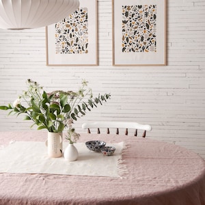 May include: A white brick wall with two framed prints of abstract patterns in shades of brown, grey, and yellow. A white pendant light hangs from the ceiling above a pink tablecloth with a white runner and two small bowls with blue and white patterns.