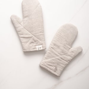 May include: Two beige linen oven mitts with a small white label on one mitt. The mitts are quilted and have a hanging loop.