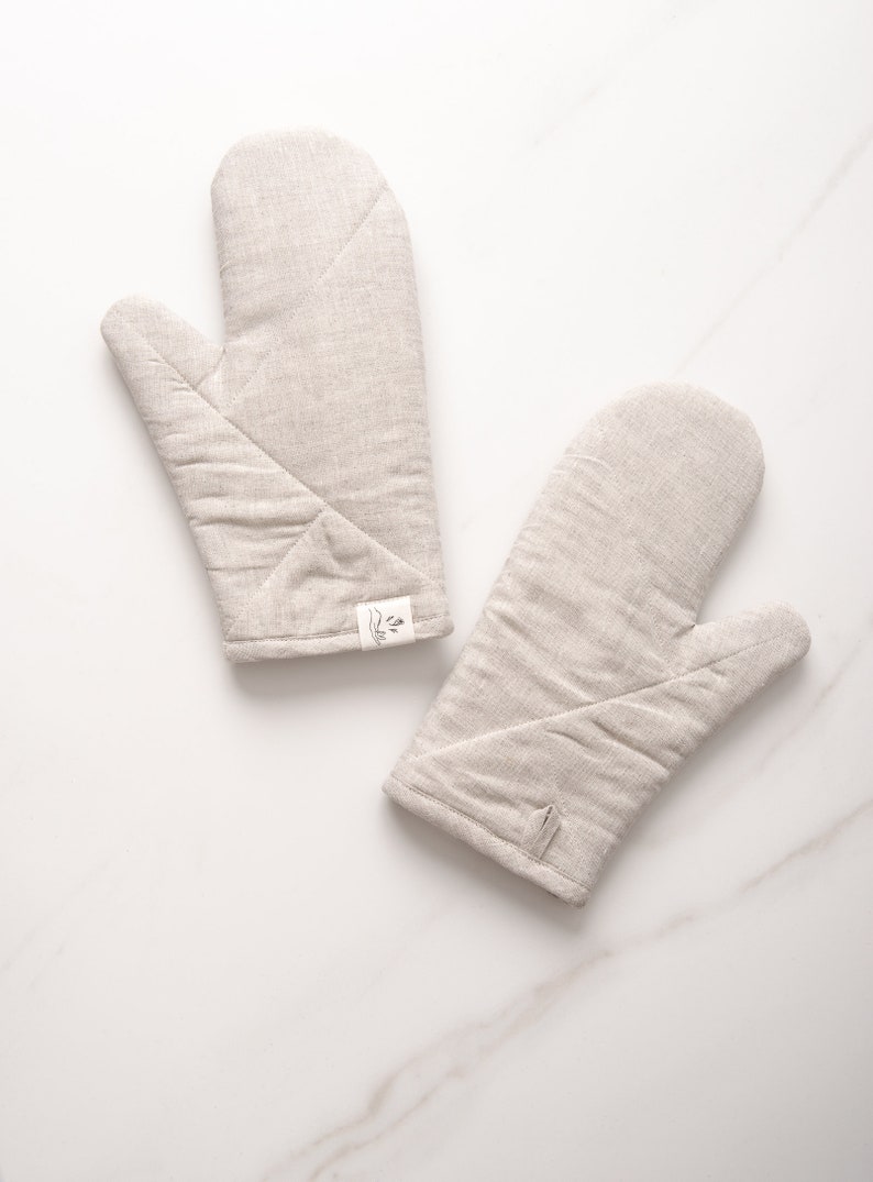 May include: Two beige linen oven mitts with a small white label on one mitt. The mitts are quilted and have a hanging loop.