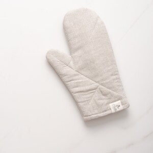 May include: A beige linen oven mitt with a small white label that says "The Little Market" with a simple line drawing of a plant.