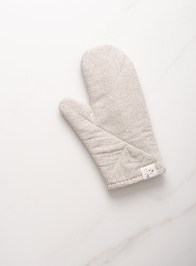 May include: A beige linen oven mitt with a small white label that says "The Little Market" with a simple line drawing of a plant.